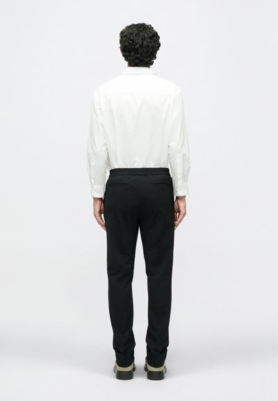 Balta ilgomis rankovėmis marškiniai, juodos kelnės su elastinga juostele ir juodi batai su žaliais akcentais, matomi iš nugaros. Paprastas ir pritaikytas dizainas.