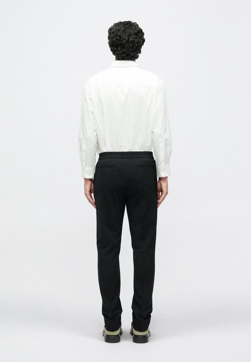 Witte blouse met lange mouwen, zwarte broek met elastische taille, en zwarte schoenen met groene accenten, gezien vanuit de achterkant. Eenvoudig en verzorgd ontwerp.