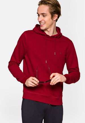Bordowy hoodie wykonany z miękkiego materiału, z kieszenią kangurzą, sznurkami do regulacji oraz małym logo na klatce piersiowej. Model trzyma okulary.