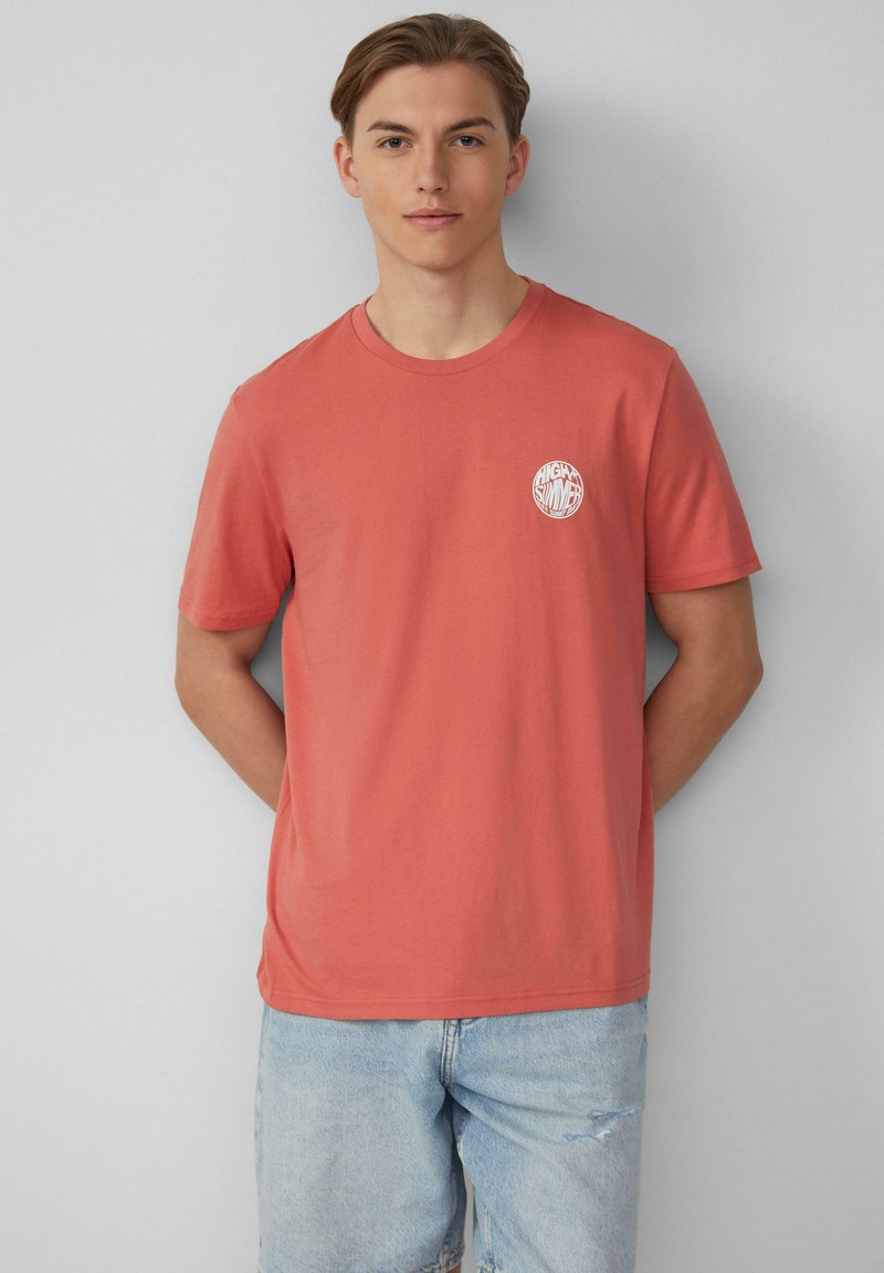 T-shirt en coton corail avec un col rond et des manches courtes ; l'avant à gauche présente un logo circulaire blanc avec du texte. Coupe décontractée.