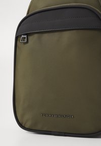 Olive green backpack with a smooth textile finish, featuring a black top zip pocket and embossed logo at the bottom.
