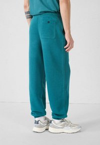 Teal katoenen joggers met een elastische tailleband, geknoopte boorden en een achterzak met een enkele knoopdetail, gecombineerd met lichte sneakers.