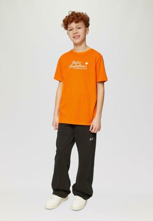 Pomarańczowy bawełniany t-shirt z białym tekstem logo, w połączeniu z czarnymi luźnymi spodniami i białymi trampkami, stojący na neutralnym tle.