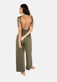 Olive green, backless jumpsuit with adjustable tie straps, elastic waistband, and wide-leg design; fabric appears textured and lightweight.