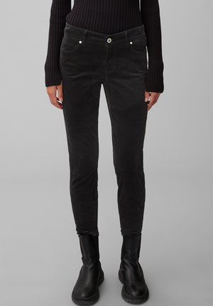 Frau trägt eng anliegende schwarze Cordhose, schwarzen gerippten Pullover und schwarze Stiefeletten, steht vor einem grauen Hintergrund.