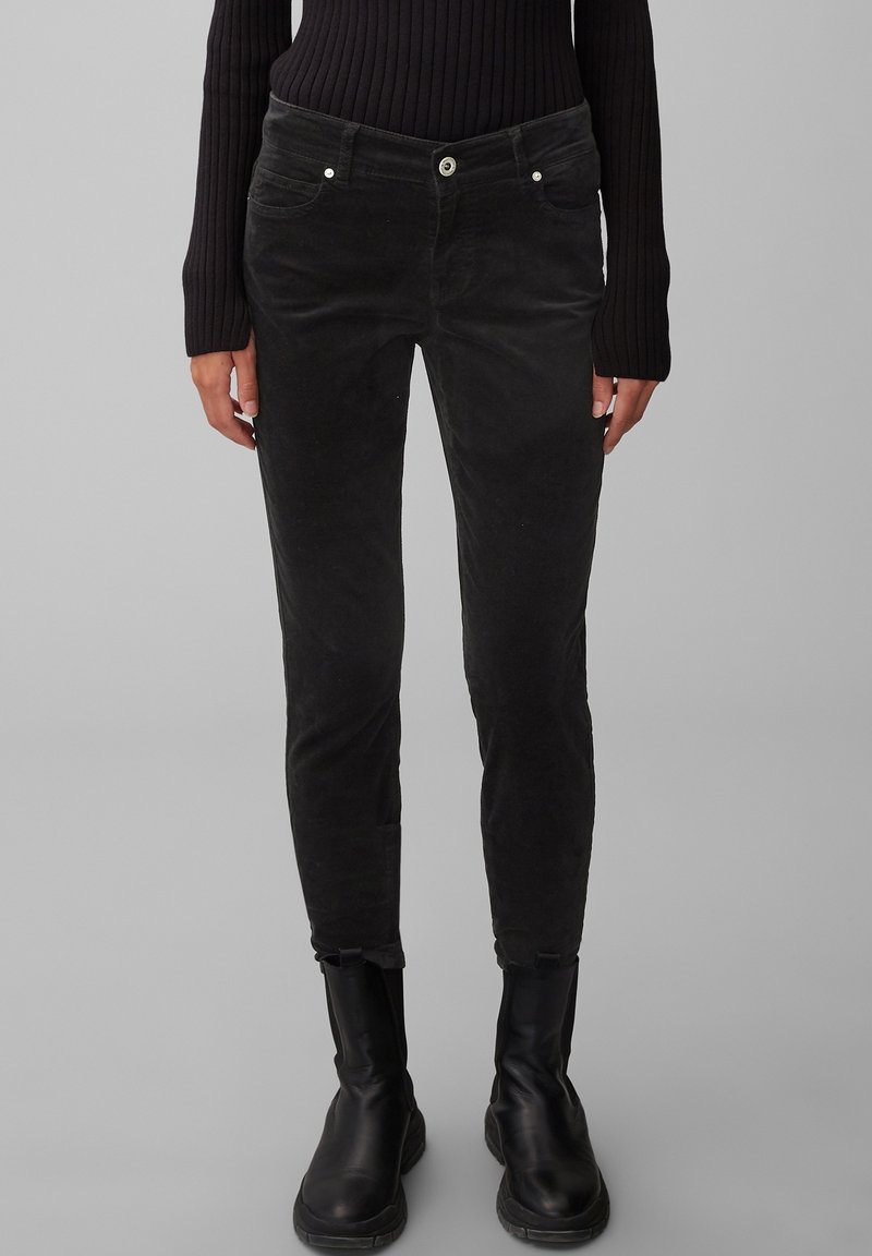 Femme portant un pantalon slim en velours côtelé noir, un pull côtelé noir et des bottines noires, debout devant un fond gris.