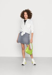 Witte blouse met knoop aan de voorkant, grijze denimrok en felgroene handtas. Sportieve schoenen met pastelaccenten maken de look compleet.