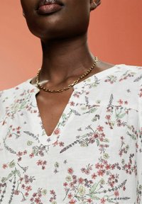 Personne à la peau foncée portant un chemisier blanc avec de petits motifs floraux roses et verts et un collier chaîne en or sur un fond pêche.