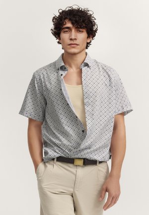 Junger Mann mit lockigem Haar trägt ein gemustertes kurzärmliges Hemd über einem beigen Tanktop, beigen Hosen und einem dunklen Gürtel mit goldener Schnalle.