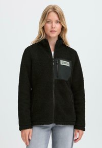 Black fleece jacket with a zip front, high collar, and a patch pocket featuring a label on the left side. Soft, textured fabric.