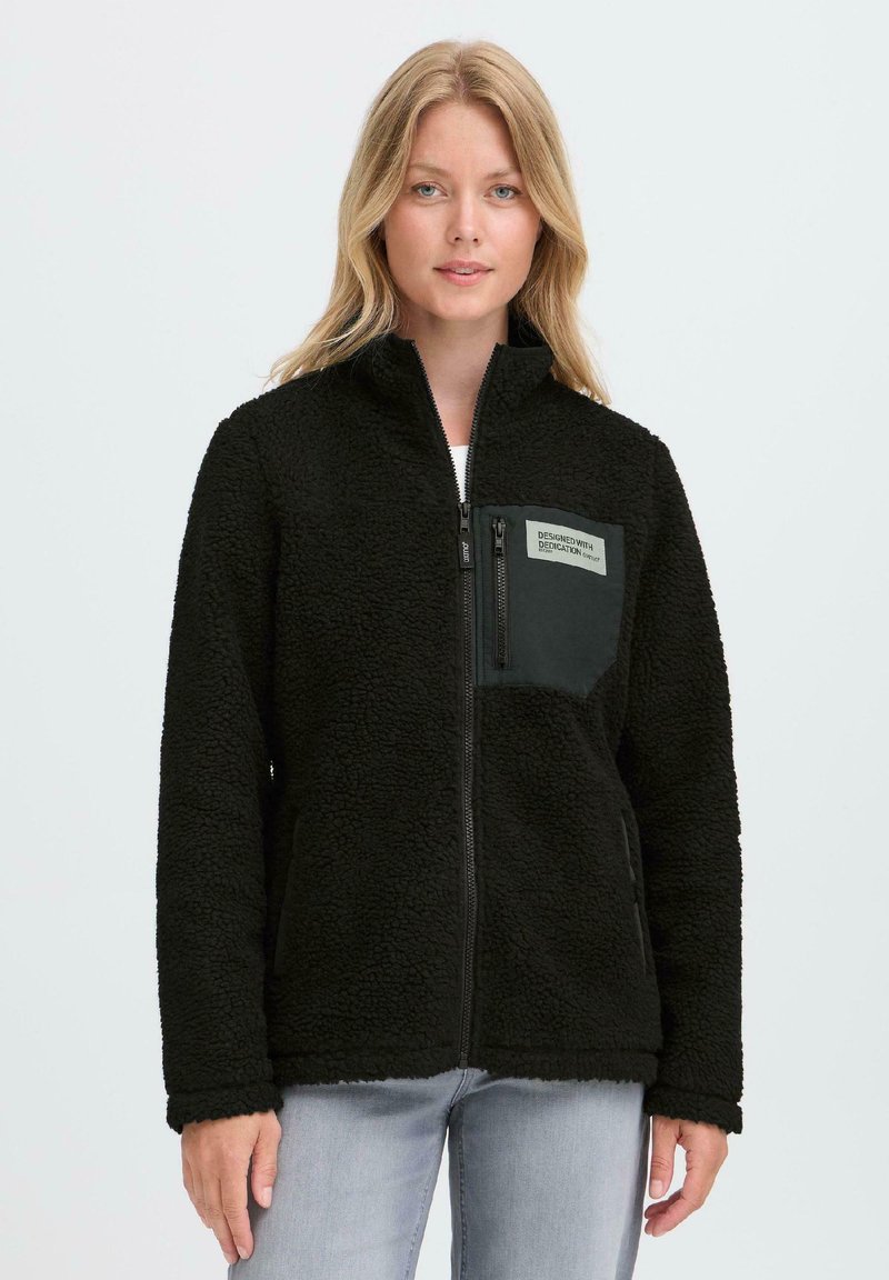Black fleece jacket with a zip front, high collar, and a patch pocket featuring a label on the left side. Soft, textured fabric.