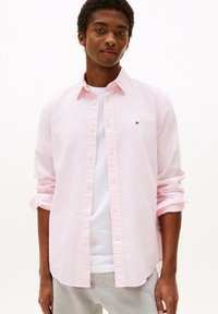 Helles rosa Hemd mit Knopfleiste und strukturiertem Muster, hochgekrempelten Ärmeln und einem kleinen Logo auf der Brust, getragen über einem weißen T-Shirt.