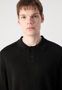 Schwarzes Polohemd mit Kragen, ausgestattet mit drei schwarzen Knöpfen und einem strukturierten Stoff. Subtiles Branding auf der Brust; minimalistisches Design.