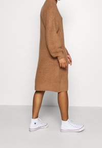 Abito maglione lungo a maniche lunghe in marrone con texture a coste, indossato con sneakers bianche alte con accenti rossi e blu.