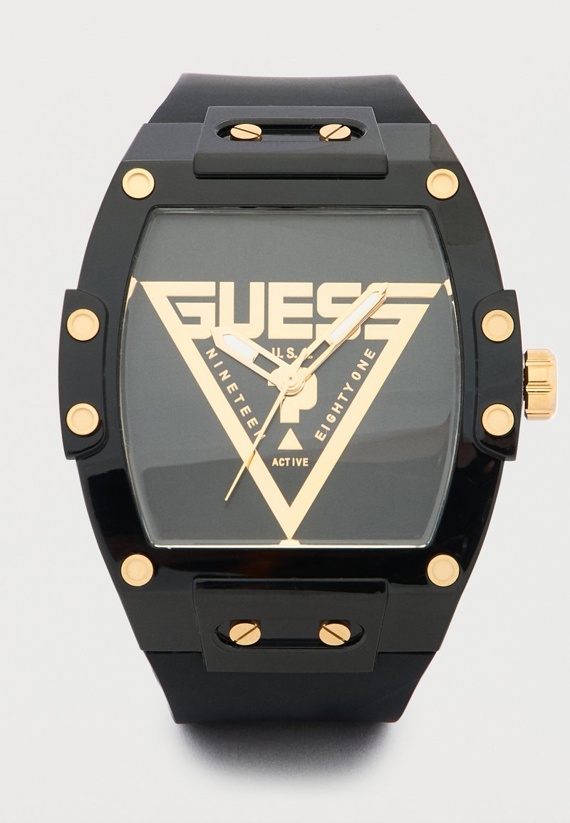 Montre-bracelet Guess noire avec des vis en or, des aiguilles et un logo triangulaire sur le cadran affichant l'heure 10:10:37.