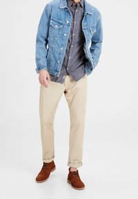 Leren jack in lichtblauw, dichtgeknoopt, met twee borstzakken; grijze overhemd eronder; beige broek; bruine suède schoenen.