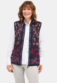 Gebreide vest met een marineblauwe achtergrond en bloemen- en paisleymotieven in rood, groen en wit. Voorzien van een ritssluiting, gecombineerd met een witte button-up shirt.
