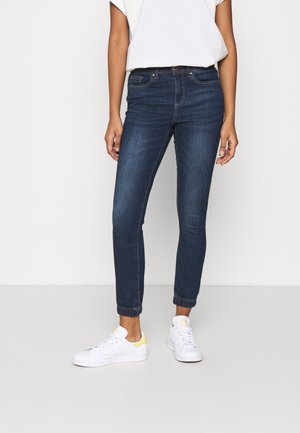 Persoon draagt donkerblauwe slim-fit jeans, witte sneakers met gele accenten en een wit kortärmelig shirt, staand tegen een effen achtergrond.
