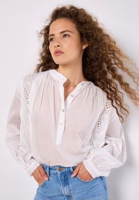 Weiße Baumwollbluse mit Knopfleiste, bestickten Akzenten und langen, voluminösen Ärmeln. Verfügt über eine strukturierte Detail entlang der Schultern.