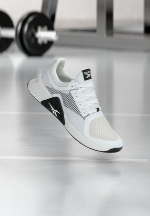 Biało-czarny but sportowy Reebok unoszący się nad podłogą siłowni, w tle rozmyte hantle i sprzęt.