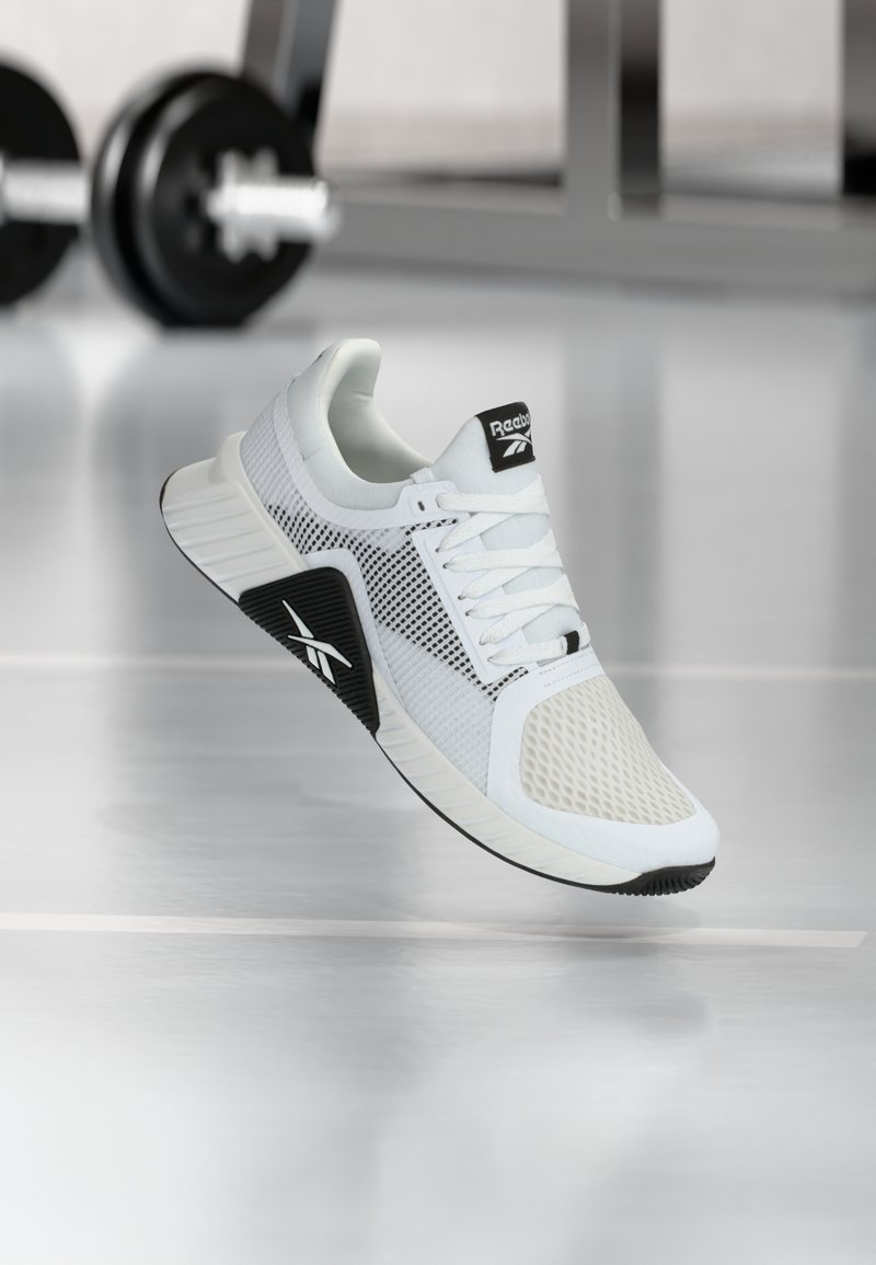 Λευκό και μαύρο αθλητικό παπούτσι Reebok που αιωρείται πάνω από το πάτωμα ενός γυμναστηρίου με θαμπά αλτήρες και εξοπλισμό στο φόντο.