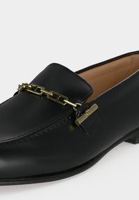 Mocassin en cuir noir avec chaîne carrée dorée et détail barre sur le dessus, doublure intérieure couleur fauve et petit talon.