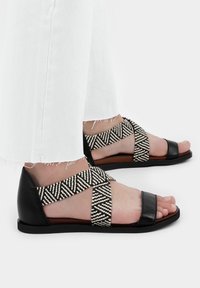 Černé kožené sandály s tkanými páskami s černobílým vzorem. Design s otevřenou špičkou, plochá podrážka a odpočívají na bílých džínových kalhotách.