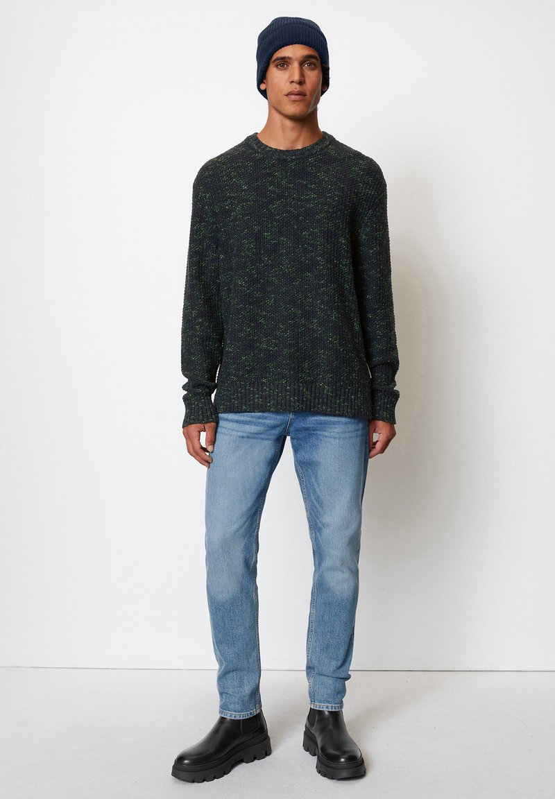 Pull en tricot vert foncé avec une finition texturée, associé à un jean bleu clair et des bottines noires. Le modèle porte un bonnet bleu marine.