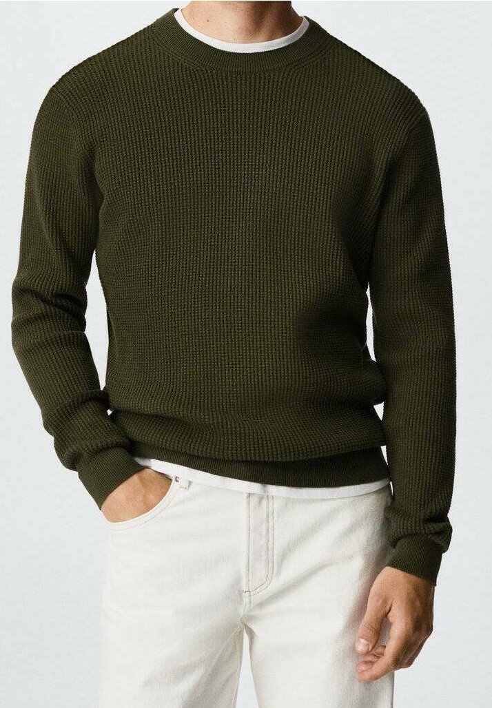 Pull en tricot vert olive avec un col rond, une texture côtelée et des poignets ajustés. Associé à un pantalon de couleur claire.