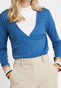 Blå ribbad cardigan med V-ringning och knäppning, ihop med beige byxor med randiga detaljer vid ärmsluten.