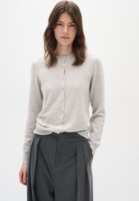 Vaalea harmaa neulecardigan, jossa on pyöreä kaula-aukko, napitettava etuosa, joustavat hihansuut ja helma. Yllä tummat räätälöidyt housut.
