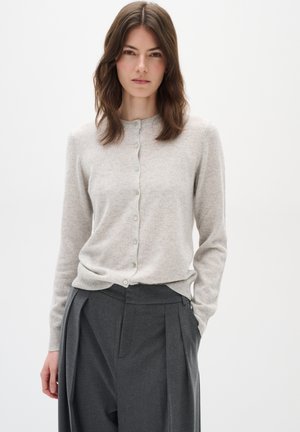 Cardigan lavorato a maglia grigio chiaro con scollo rotondo, chiusura a bottoni davanti, polsini e orlo a coste. Indossato sopra pantaloni scuri su misura.