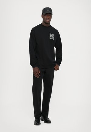 Mann in schwarzem Outfit mit Kappe, langärmligem Shirt mit weißem Text auf der Brust, schwarzen Hosen und schwarzen Schuhen, der vor einem weißen Hintergrund steht.