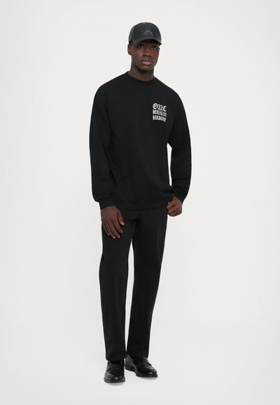 Mann in schwarzem Outfit mit Kappe, langärmligem Shirt mit weißem Text auf der Brust, schwarzen Hosen und schwarzen Schuhen, der vor einem weißen Hintergrund steht.