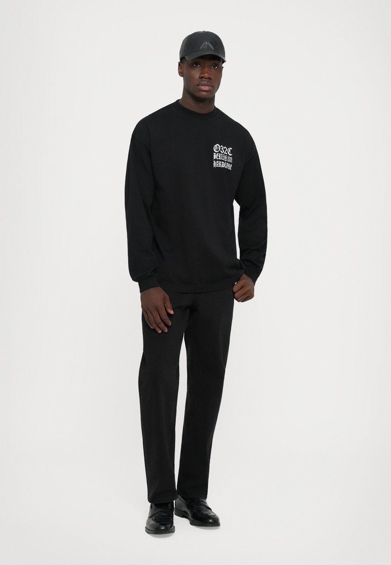 Mann in schwarzem Outfit mit Kappe, langärmligem Shirt mit weißem Text auf der Brust, schwarzen Hosen und schwarzen Schuhen, der vor einem weißen Hintergrund steht.