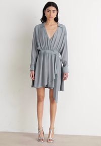 Robe portefeuille gris clair avec manches longues, ceinture à nouer et jupe fluide. Fabriquée dans un tissu doux avec un léger éclat. Sandales argentées à lanières.
