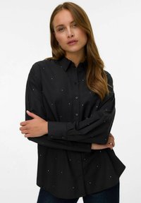 Camicia nera con bottoni, realizzata in tessuto liscio, con piccole decorazioni metalliche sparse in tutto il capo, dotata di maniche lunghe e una vestibilità rilassata.