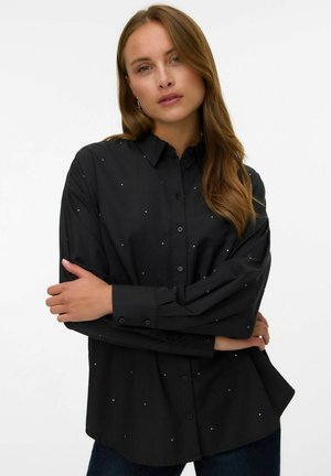 Camicia nera con bottoni, realizzata in tessuto liscio, con piccole decorazioni metalliche sparse in tutto il capo, dotata di maniche lunghe e una vestibilità rilassata.