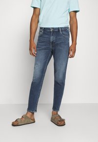 Person klädd i blå avsmalnande jeans, ljusblå t-shirt och beige mockasandaler, stående mot en enkel ljus bakgrund.