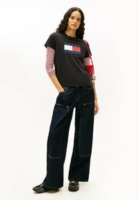 Czarna koszulka z logo "TOMMY JEANS", nałożona na białą koszulę z czerwono-białymi paskami. W zestawieniu z luźnymi, ciemnoniebieskimi dżinsami i czarnymi butami.