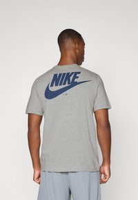 Pilkos spalvos medvilninė marškinė su apvaliu kaklu, su dideliu tamsiai mėlynu „Nike“ logotipu ant nugaros, trumpomis rankovėmis ir laisvu siluetu.