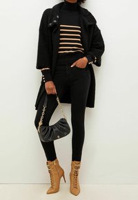 Cappotto teddy nero, maglione a righe nero e marrone, jeans skinny neri, stivali con tacco a stiletto color marrone chiaro con lacci e borsa nera con dettagli in catena dorata.