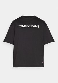 Camiseta negra de manga corta hecha de algodón, con un logo blanco "TOMMY JEANS" en la parte frontal y un pequeño parche del logo en el dobladillo.