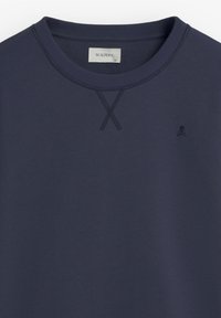 Marinblå sweatshirt med ribbad rund halsringning, korsstygnsdetalj och liten broderad dödskallelogotyp på bröstet. Mjuk textur.