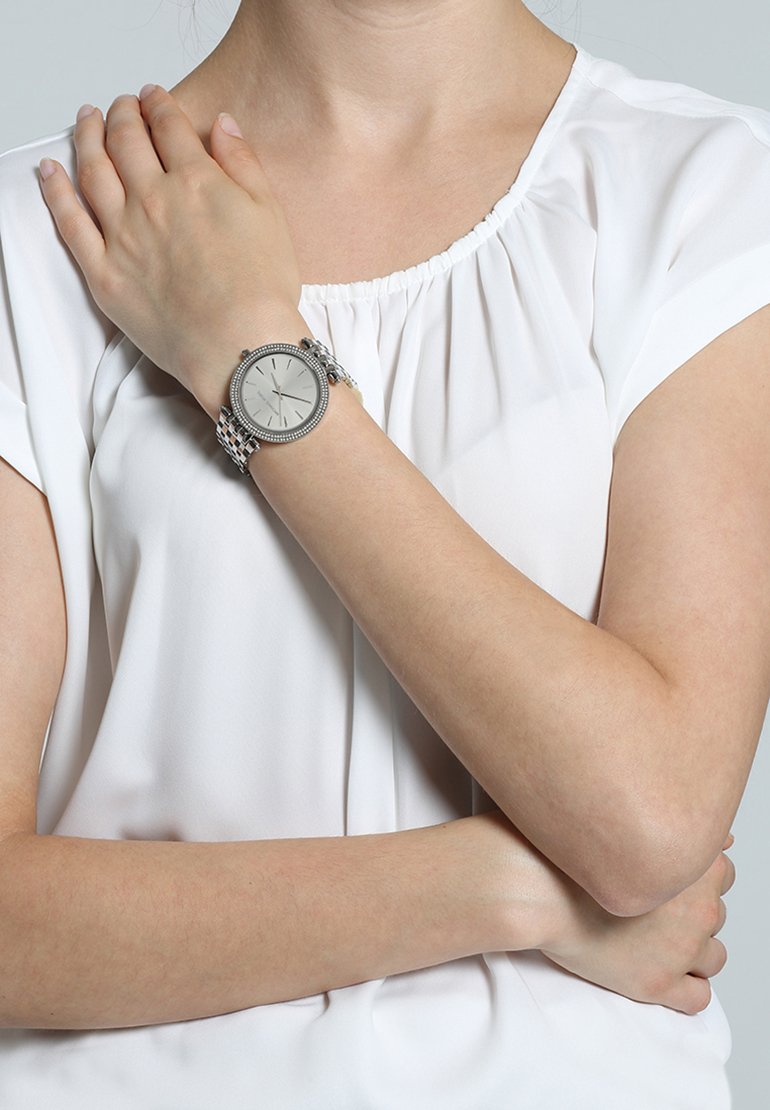 Woman wearing white short-sleeve blouse, silver wristwatch on left wrist, left hand resting on right shoulder, right arm crossed.