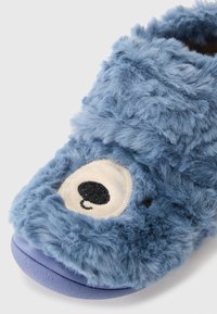 Pantufla de peluche azul con una textura suave, forma redondeada y una cara de oso decorativa con detalles de ojos color crema y costuras negras.