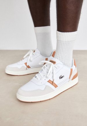 Jambes à la peau foncée portant des chaussettes blanches côtelées et des baskets blanches Lacoste avec des touches beige clair sur un sol beige lisse.
