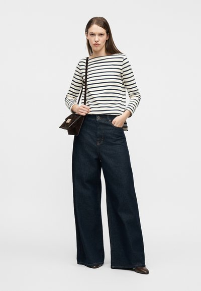 Γυναίκα που στέκεται με το ένα χέρι στην τσέπη, φοράει λευκό και ναυτικό μπλε ριγέ μπλούζα με μακριά μανίκια, φαρδιά σκούρα τζιν παντελόνια, καφέ παπούτσια και καφέ τσάντα ώμου.