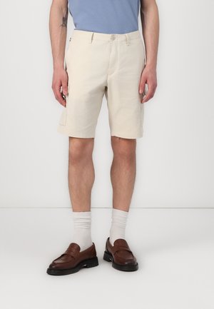 Homme portant un short cargo crème, des chaussettes blanches, des mocassins marron et une chemise bleu clair, debout devant un fond blanc.
