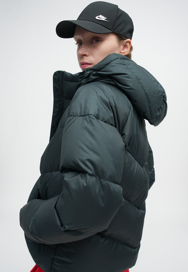 Dark green puffer jacket with hood, featuring quilted sections, front zipper, and side pockets. Paired with a black cap bearing a logo.